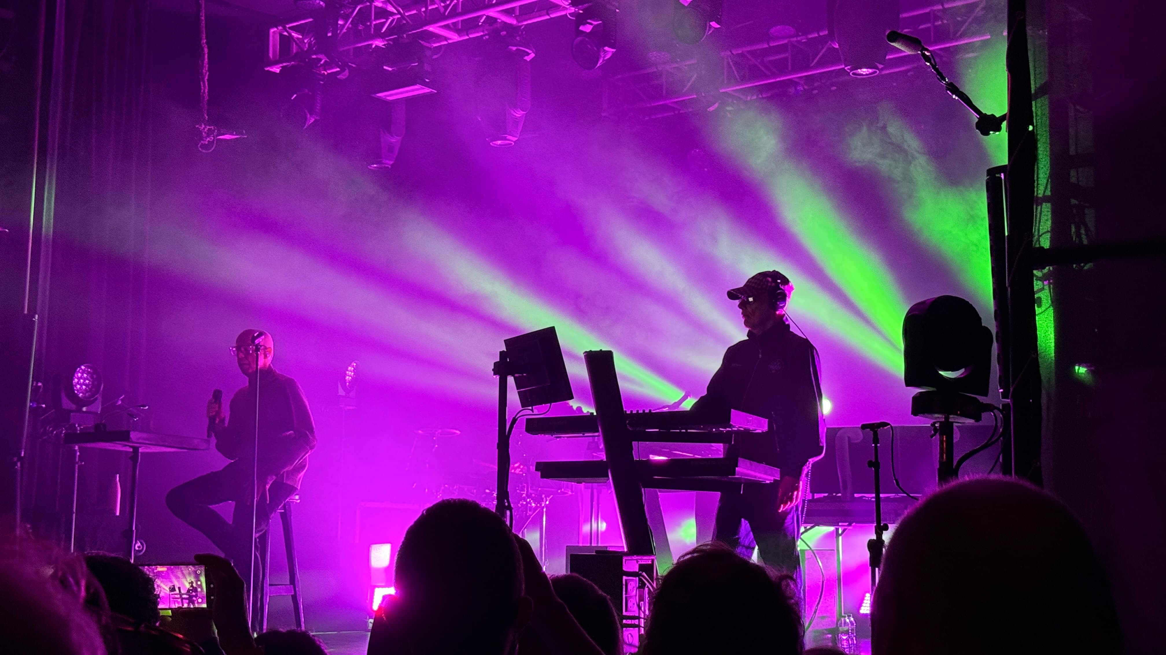 Neil Tennant and Chris Lowe from the Pet Shop Boys on stage bathed in green and purple light, Neil is sat on a stool and Chris is playing keyboards